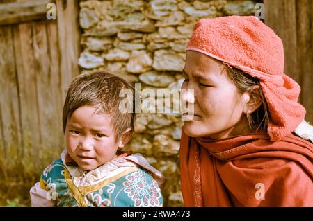 Beni, Nepal, um Mai 2012: Foto einer Mutter in rotem Schal und Kopftuch, die in Beni neben ihrer kleinen Tochter kniet, mit braunen Haaren und braunen Augen Stockfoto