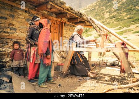 Dolpo, Nepal, um Juni 2012: Eine alte grauhaarige Frau in braunem Kleid webt auf einem Holzwebstuhl vor ihrem Steinhaus und junge Mädchen beobachten sie in Dolpo Stockfoto