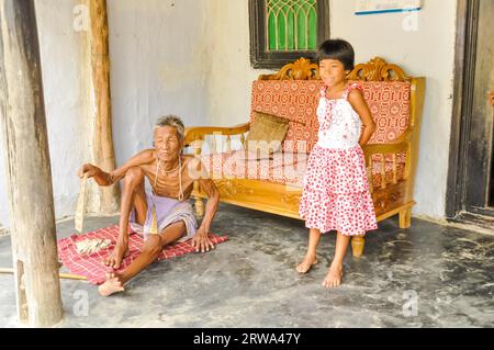 Srimongal, Bangladesch, um Juli 2012: Der alte dünne Mann sitzt auf dem Boden und seine Enkelin in rot-weißem Kleid steht in Srimongal in seiner Nähe Stockfoto