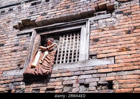 Bhaktapur, Nepal, um Juni 2012: Das junge braunhaarige Mädchen schaut aus einem Holzfenster in einem alten Backsteinhaus in Bhaktapur, Nepal, auf die Fotokamera herunter. Stockfoto