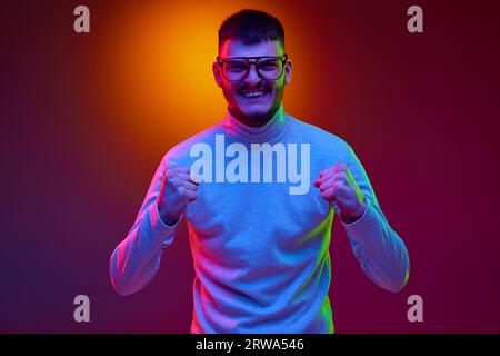 Hübscher junger bärtiger Mann, der lässig in die Kamera schaut, mit geballten Fäusten im Hintergrund in gemischten Farben, Licht, Filter. Stockfoto