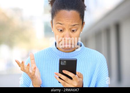 Porträt einer verblüfften schwarzen Frau, die ihr Handy auf der Straße überprüft Stockfoto