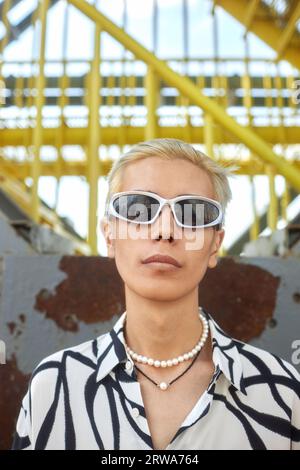 Nahaufnahme Modeporträt eines jungen asiatischen Mannes, der eine Sonnenbrille trägt und die Kamera vor der urbanen Metallstruktur betrachtet Stockfoto