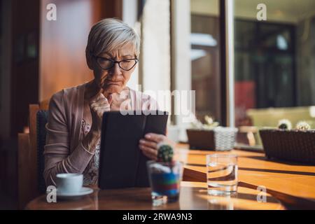Reife Frau sitzt im Café und benutzt ein digitales Tablet. Stockfoto