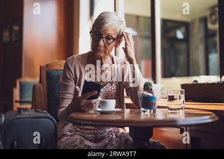 Reife Frau sitzt in einem Café am Flughafen und wartet auf das Boarding. Sie prüft den Zeitplan auf dem Smartphone. Stockfoto
