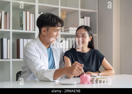 Glückliches asiatisches junges Paar liebt die Hand, die Münze in eine Spardose steckt, um Geld zu sparen. Unternehmensfinanzierung, Einlage beim Bankgeschäft für die gemeinsame Finanzplanung Stockfoto