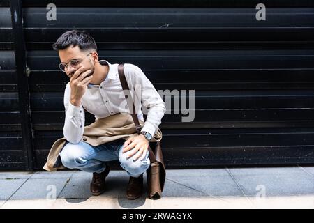 Junger, gestresster und depressiver Geschäftsmann hockt vor dem Bürogebäude und wartet auf die Ergebnisse des Vorstellungsgesprächs. Der arbeitende Mann fühlt sich nervös und ängstlich Stockfoto