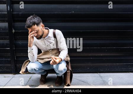 Junger, gestresster und depressiver Geschäftsmann hockt vor dem Bürogebäude und wartet auf die Ergebnisse des Vorstellungsgesprächs. Der arbeitende Mann fühlt sich nervös und ängstlich Stockfoto