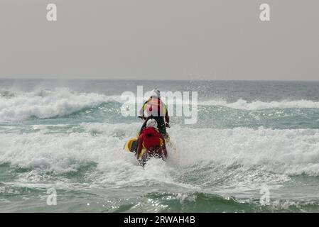 RNLI-Rettungsschwimmer auf Jet-Ski mit einem anderen auf einem Rettungsschlitten, der vor Perranporth Beach, Cornwall, UK, in raues Meer fährt. Stockfoto