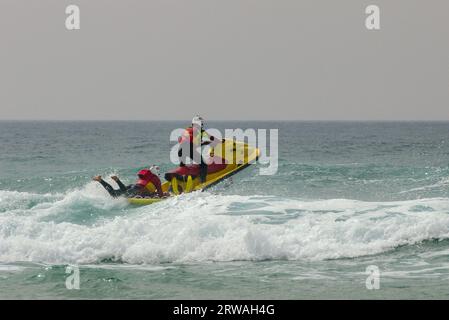 RNLI-Rettungsschwimmer auf Jet-Ski mit einem anderen auf einem Rettungsschlitten, der vor Perranporth Beach, Cornwall, UK, in raues Meer fährt. Stockfoto