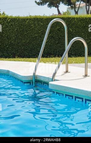 Swimmingpool-Bordsteinkante mit Leiter, mit kühl blau aussehendem Wasser und einem Buschzaun im Hintergrund. Im Sommer gibt es grünes Gras. Stockfoto