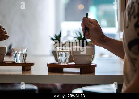 Eine Hand, die eine Tasse Kaffee umrührt, serviert in einer weißen Tasse auf einem Holztablett, mit einem Glas Wasser. Stockfoto