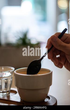 Eine Hand, die eine Tasse Kaffee umrührt, serviert in einer weißen Tasse auf einem Holztablett, mit einem Glas Wasser. Stockfoto