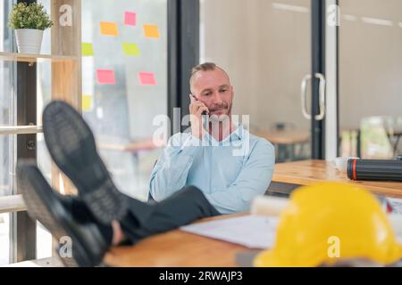 Kaukasischer Ingenieur legt Beine auf den Tisch und spricht mit Kunden im Büro auf dem Mobiltelefon Stockfoto