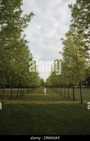 Seitenansicht einer Frau in einem weißen Hosenanzug, die auf einem Feld zwischen zwei Baumreihen steht, Weißrussland Stockfoto