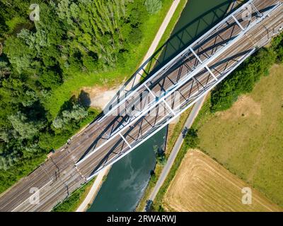 Eine beeindruckende Perspektive aus der Luft einer Eisenbahnbrücke aus Stahl, die sich elegant über einen ruhigen Fluss oder Kanal erstreckt. Das komplizierte Design und das technische Wunder dieser Brücke bilden einen markanten Kontrast zu den ruhigen Gewässern unter Ihnen. Entdecken Sie die harmonische Mischung aus Industrie und Natur in diesem fesselnden Foto. Steel Railroad Bridge Über Ruhiges Wasser: Blick Aus Der Luft. Hochwertige Fotos Stockfoto