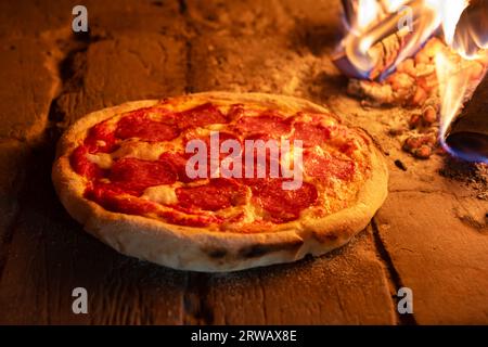 Italienische Pizza-Peperoni im Holzofen. Pizza mit Salami im heißen Ofen in der Nähe brennender Brennhölzer aus nächster Nähe. Lebensmittelfotografie Stockfoto