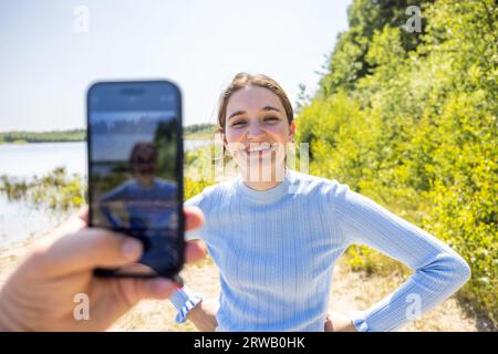 Erleben Sie die authentische Verbindung, während ein Mann ein Smartphone hält, um einen offenen Moment seiner attraktiven tausendjährigen Freundin im Herzen des Sommers festzuhalten. Ihre Liebesgeschichte entfaltet sich vor dem Hintergrund einer wunderschönen Naturkulisse. Aufrichtige Liebe: Gemeinsam Bliss Im Sommer Einfangen. Hochwertige Fotos Stockfoto