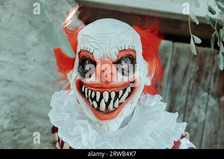 Ein verrückter böse Rothaarige Clown, der ein Kostüm mit einem weißen Fell trägt, starrt den Betrachter mit einem gruseligen Lächeln draußen vor der rustikalen Tür eines o an Stockfoto