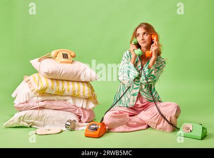 Weckrufe. Das Portrait der unzufriedenen jungen Frau im Pyjama sprach mit zwei Retro-Telefonen, die auf pastellgrünem Hintergrund isoliert waren. Stockfoto
