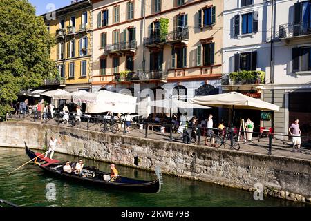 Ein Gondolier fährt durch einen Kanal in Navigli Mailand, einer der wenigen noch vorhandenen Wasserstraßen, während die Menschen in der Sommersonne speisen und eine Mahlzeit genießen. Stockfoto