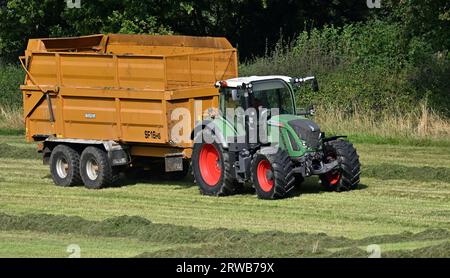 Ein großer grüner Traktor mit einem leeren gelben Anhänger, der bereit ist, das Gras von einem Feld zur Lagerung zu bewegen. Stockfoto