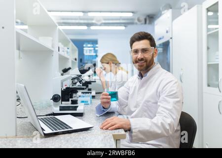 Porträt eines lächelnden jungen männlichen Wissenschaftlers, Chemiker, der in einem Labor arbeitet und an einem Tisch sitzt, der eine Flasche mit einer blauen Flüssigkeit in den Händen hält. Lächelnd in die Kamera. Stockfoto