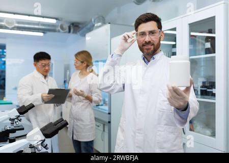 Porträt eines lächelnden jungen Apothekers, der in einem Labor in Uniform steht und ein Plastikgefäß mit Material in den Händen hält. Zeigt das Ergebnis der Kamera an. Die Kollegen diskutieren dahinter. Stockfoto