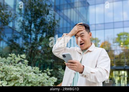 Schockierter junger asiatischer Mann, der draußen auf der Straße stand und auf den Bildschirm schaute und seinen Kopf überrascht hielt. Stockfoto