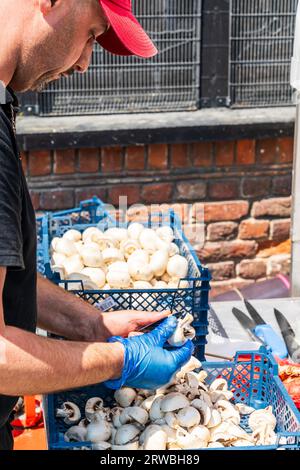 Nahaufnahme eines Mannes im Freien, der Pilze über ein großes Plastiktablett mit geschnittenen Pilzen mit einem anderen Tablett neben ihm mit ungeschnittenen Pilzen schneidet. Stockfoto