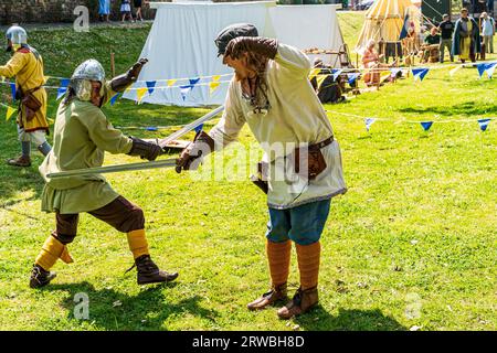 Zwei Männer in historischen Kostümen, die mit Schwertern im Sonnenschein während einer mittelalterlichen Nachstellung in einem von gelben und blauen Flaggen umgebenen Gebiet jagen Stockfoto