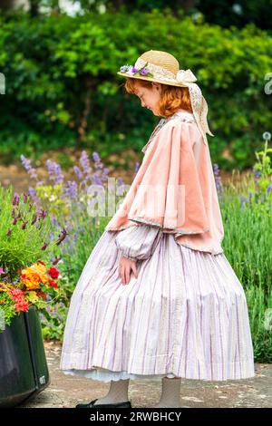 Junge Teenager-Frau im Victoria-Kostüm, die traurig auf einige Blumen in einer Plastikschale in einem Garten schaut. Sie trägt einen Strohhut, ein Tuch und einen weiten Rock. Stockfoto