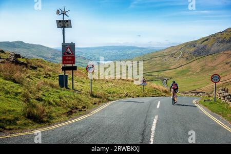 Radfahrer, die den Kampf erklimmen und sich der Spitze nähern, mit dem 20 %-Gradienten-Schild in Sichtweite, in der Nähe von Ambleside im Lake District, Cumbria, England, Großbritannien Stockfoto