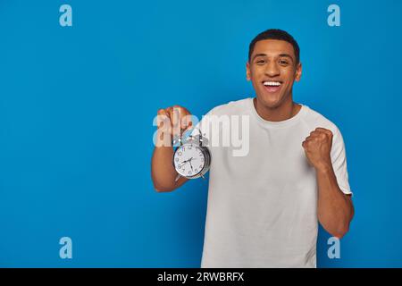 Aufgeregter afroamerikanischer Mann mit offenem Mund, der Wecker auf blauem Hintergrund hält, Gesichtsausdruck Stockfoto