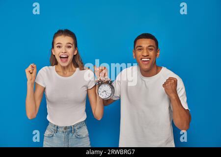 Aufgeregtes interrassisches Paar mit offenem Mund, der Wecker auf blauem Hintergrund hält, Gesichtsausdruck Stockfoto