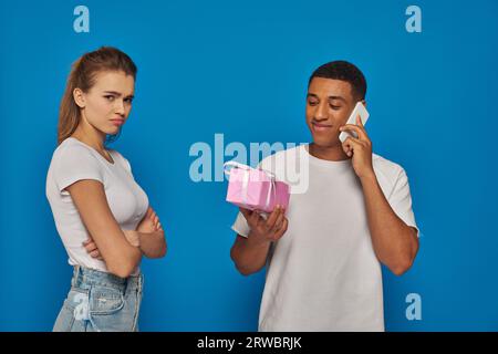 Glücklicher afroamerikanischer Mann, der telefoniert und ein Geschenk in der Nähe einer beleidigten Frau auf blauem Hintergrund hält Stockfoto