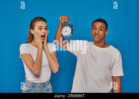 Morgenzeit, glücklicher afroamerikanischer Mann, der Wecker in der Nähe einer erstaunten Frau auf blauem Hintergrund hält Stockfoto