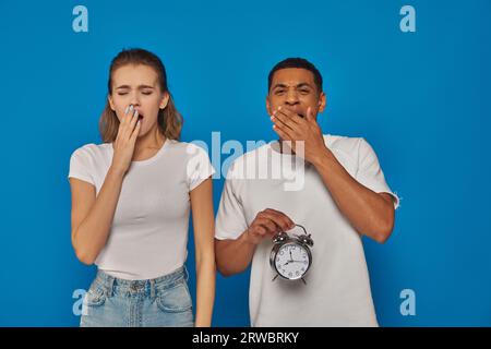 afroamerikanischer Mann mit Wecker und Gähnen mit Frau vor blauem Hintergrund, verschlafenes Paar Stockfoto