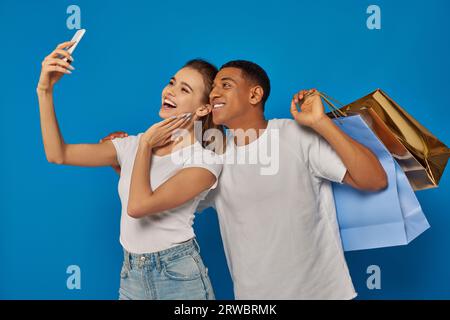 Konsumismus, glückliches, interrassisches Paar, das Einkaufstaschen hält und Selfie vor blauem Hintergrund macht Stockfoto