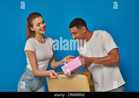 Glückliches multikulturelles Paar, das Geschenk in die Einkaufstasche auf blauem Hintergrund steckt, Konsumkonzept Stockfoto