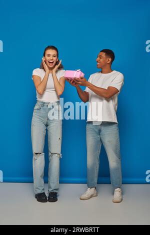 Glücklicher afroamerikanischer Mann, der ein Geschenk an eine aufgeregte Freundin vor blauem Hintergrund präsentiert Stockfoto