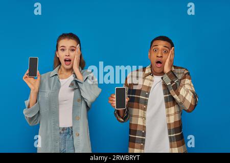 Schockierte Frau und staunte afroamerikaner mit Smartphones mit leerem Bildschirm vor blauem Hintergrund Stockfoto
