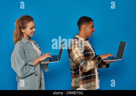 Glückliche interrassische Freiberufler mit Laptops vor blauem Hintergrund, verschiedene Kulturen, Mann und Frau, Seite Stockfoto