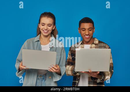 Glückliche interrassische Freiberufler mit Laptops vor blauem Hintergrund, Mann und Frau in verschiedenen Kulturen Stockfoto