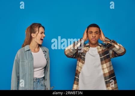 Emotionale Frau schreit in der Nähe eines afroamerikanischen Mannes, der seine Ohren auf blauem Hintergrund bedeckt, ausdrucksstark Stockfoto