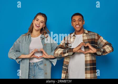 Fröhliches, multikulturelles Paar, das Herzzeichen mit Händen zeigt und die Kamera auf blauem Hintergrund betrachtet Stockfoto