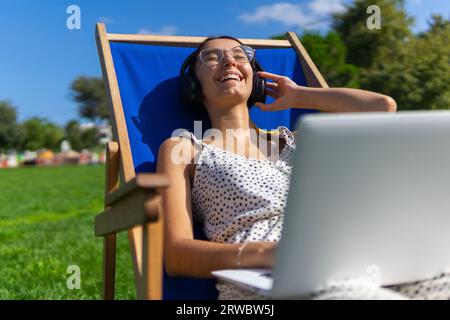 Glückliche junge Frau in einer Brille mit Laptop und Kopfhörern, die sich im Sommer auf dem Stuhl entspannen und Musik hören Stockfoto