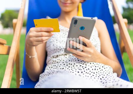 Eine anonyme Frau sitzt im Stuhl mit Kreditkarte in der Hand im Park und kauft online mit dem Handy Stockfoto