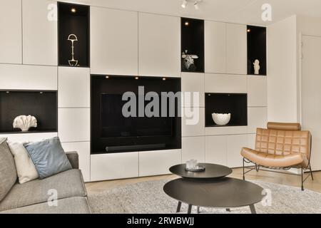Das geräumige Apartment verfügt über ein modernes Wohnzimmer mit großem Fernseher und Dekor auf schwarzen Regalen und weißen Wänden Stockfoto