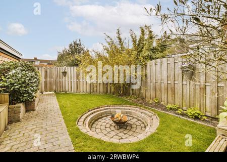Feuerstelle und Steinmauer in der Mitte des Grases und gegen Holzzaun im privaten Garten im Hinterhof gegen bewölkten Himmel Stockfoto